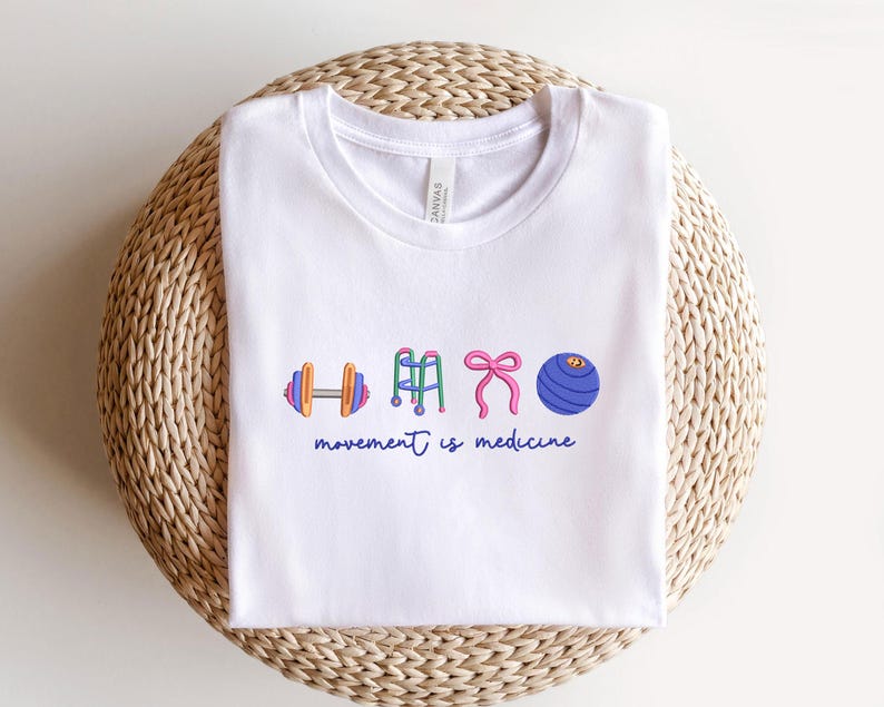 Puede incluir: Camiseta blanca con cuello redondo con ilustraciones coloridas de equipos de ejercicio: una mancuerna, un andador, un lazo rosa y una pelota de ejercicios. El texto "movement is medicine" est&aacute; escrito en azul debajo.