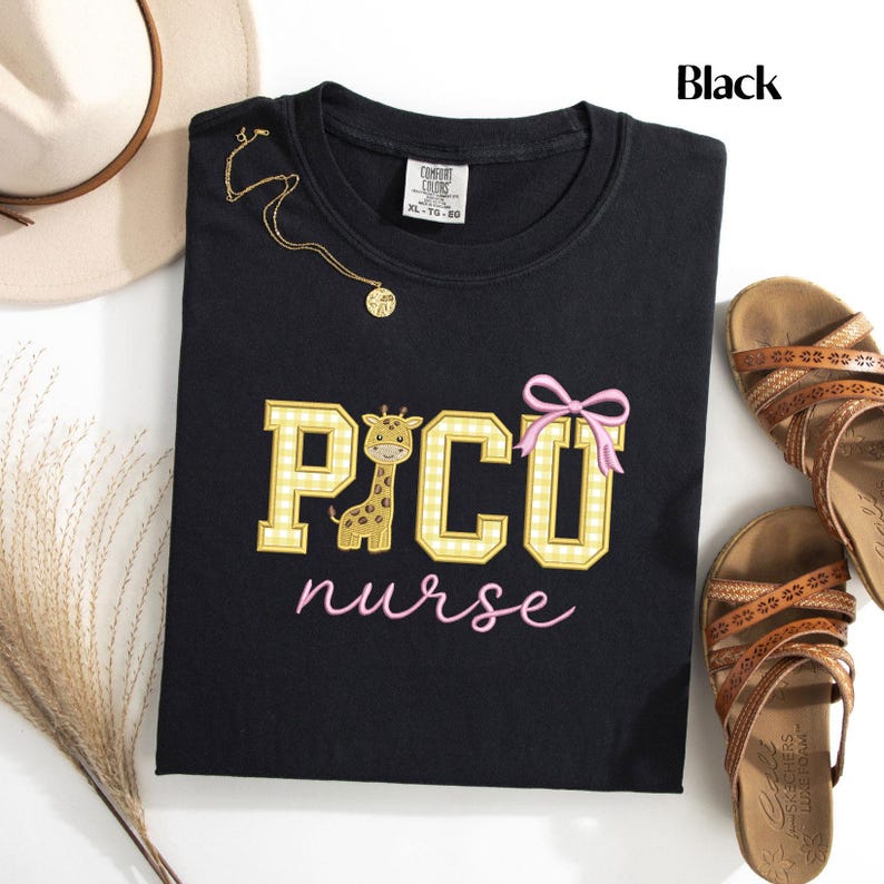 Puede incluir: Sudadera negra de cuello redondo con el texto "PICO nurse" en bordado amarillo y rosa. El dise&ntilde;o incluye una jirafa y un lazo rosa. Tambi&eacute;n se ven un collar dorado y sandalias.