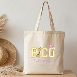 Puede incluir: Bolso tote de color crema con el texto "PICU nurse" en una fuente l&uacute;dica. Las letras son amarillas con un estampado de cuadros, y un gr&aacute;fico de jirafa est&aacute; en la letra "C". Un lazo rosa est&aacute; en el lado derecho.