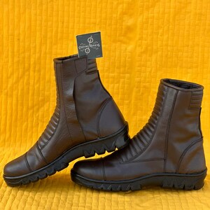 May include: A pair of brown leather boots with a ridged design on the upper shaft. The boots have a zipper closure and a black, textured sole. The boots are displayed on a yellow background.