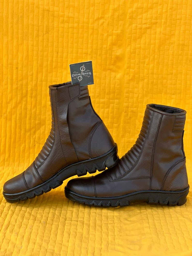 May include: A pair of brown leather boots with a ridged design on the upper shaft. The boots have a zipper closure and a black, textured sole. The boots are displayed on a yellow background.