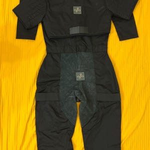 May include: Black protective suit with a quilted upper section and attached pants. The suit has a collar, short sleeves, and reinforced panels. Small square patches with a logo are visible on the chest and lower torso. The suit is laid out on a yellow surface.