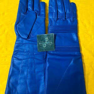 May include: A pair of long, royal blue gauntlet-style gloves featuring ribbed detailing on the knuckles and wrists. A small, square dark green tag with the text "COSTUME REPLICAS" and a circular logo is placed centrally between the two gloves.