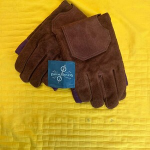 May include: A pair of brown suede gloves with a dark brown patch on the back of the hand. The gloves have purple accents at the wrist and a blue tag that reads "Costume Replicas". The gloves are displayed on a yellow textured surface.