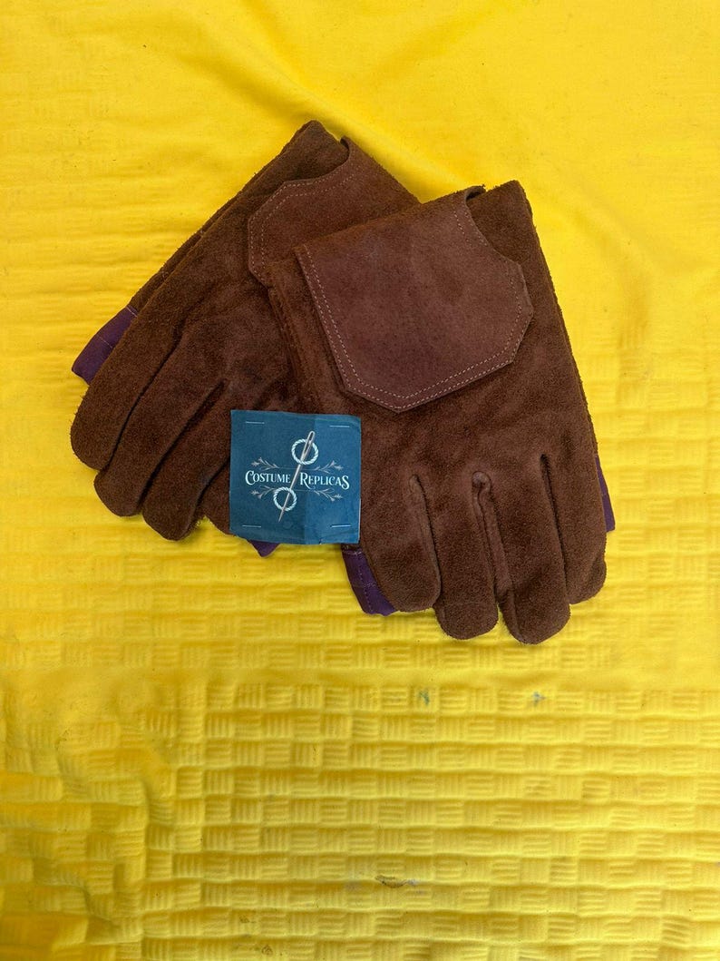May include: A pair of brown suede gloves with a dark brown patch on the back of the hand. The gloves have purple accents at the wrist and a blue tag that reads "Costume Replicas". The gloves are displayed on a yellow textured surface.