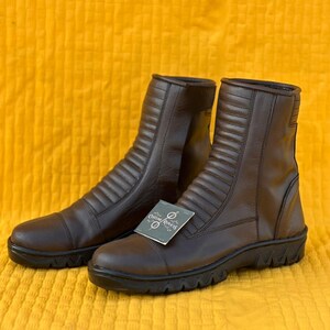 May include: A pair of brown leather boots with a ridged design on the front and sides. The boots have a round toe and a black sole. A tag that says "Custom Replcas" is attached to one of the boots.