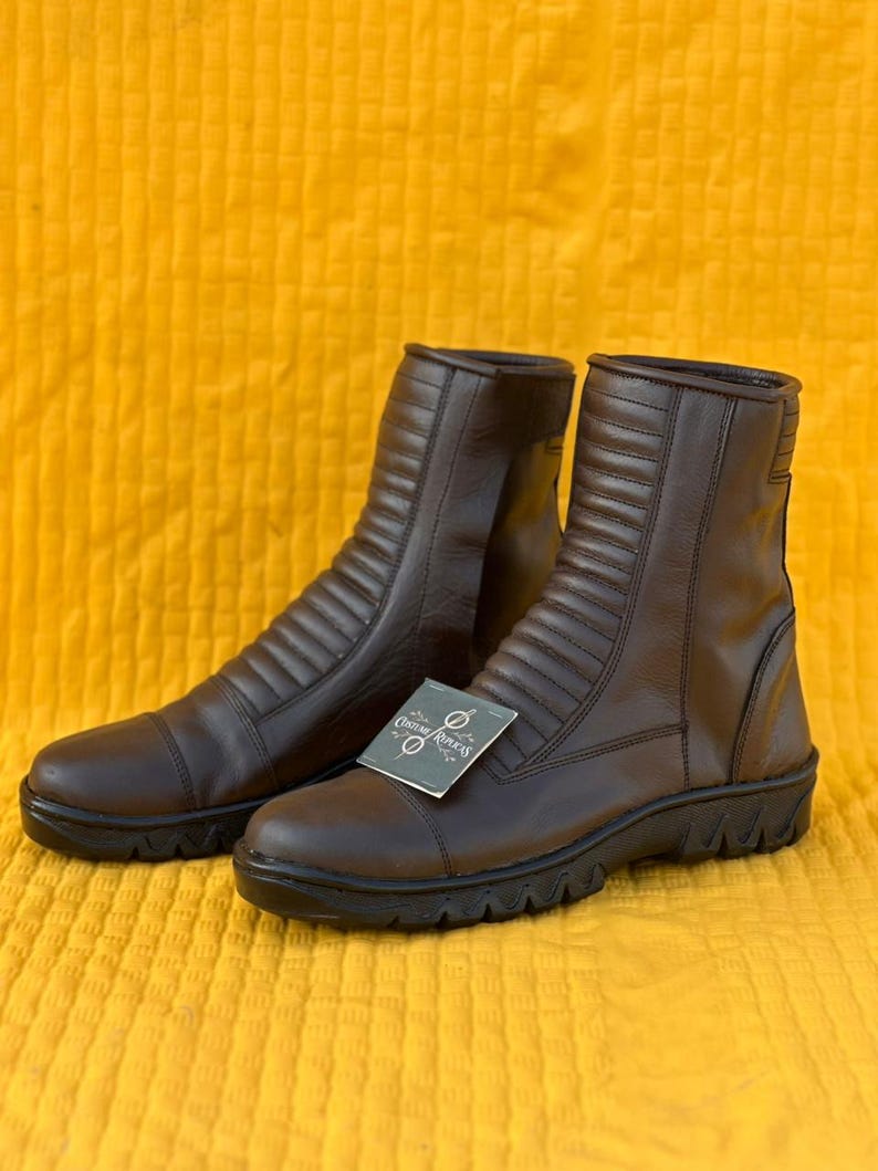 May include: A pair of brown leather boots with a ridged design on the front and sides. The boots have a round toe and a black sole. A tag that says "Custom Replcas" is attached to one of the boots.