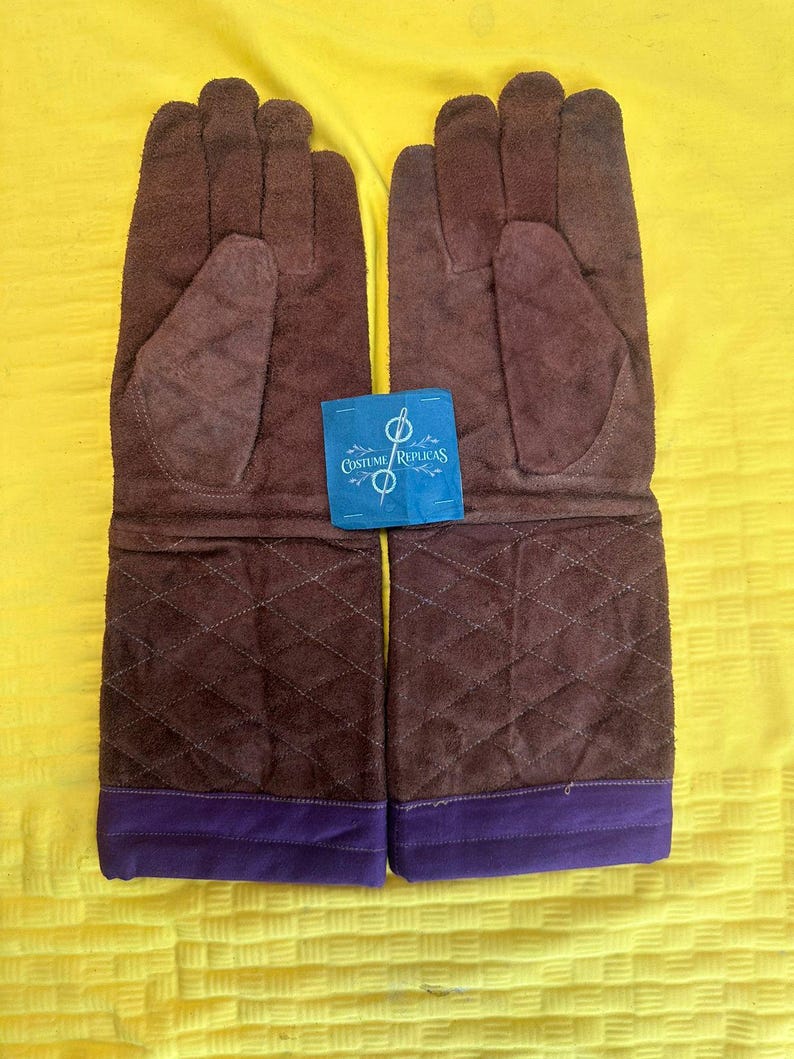 May include: A pair of brown suede gloves with a quilted pattern on the lower portion and a purple cuff. The gloves have a tag that reads "Costume Replicas".