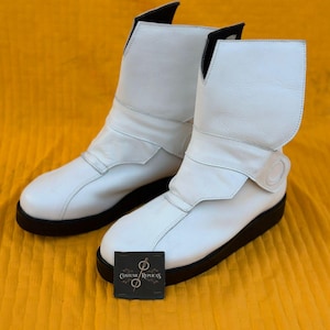 Könnte beinhalten: Ein Paar weiße Lederstiefel mit schwarzen Sohlen. Die Stiefel haben ein einzigartiges Design mit einer Klappe, die sich über die Oberseite faltet. Die Stiefel werden auf gelbem Hintergrund präsentiert. Eine kleine schwarze Karte mit weißem Text befindet sich vor den Stiefeln.