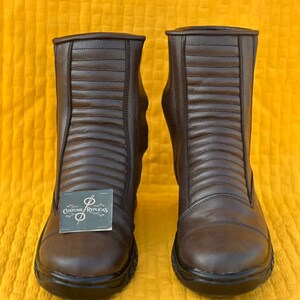 May include: A pair of brown leather boots with a ridged design on the front. The boots have a rounded toe and a black sole. A tag with the text "Costume/Replicas" is attached to one boot.