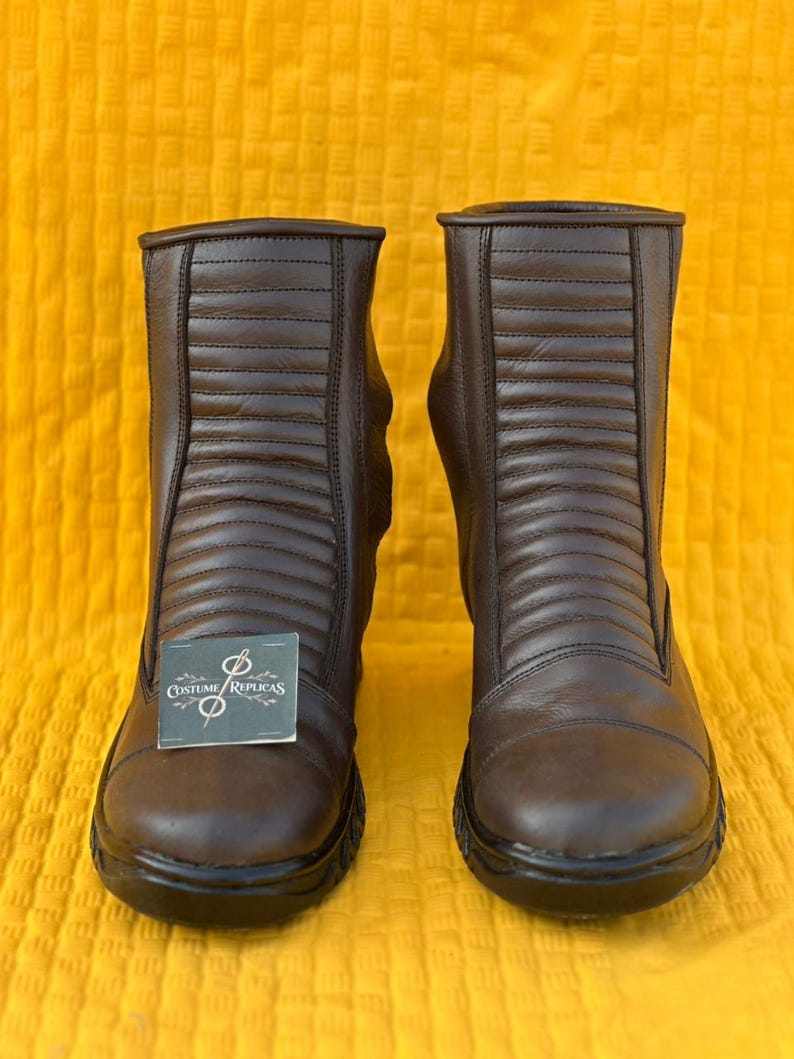 May include: A pair of brown leather boots with a ridged design on the front. The boots have a rounded toe and a black sole. A tag with the text "Costume/Replicas" is attached to one boot.