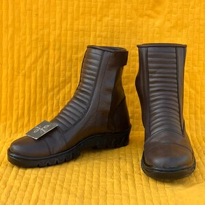 May include: A pair of brown leather ankle boots with a ridged design on the upper. The boots have a black sole and a small tag attached to one boot. The boots are displayed on a yellow textured surface.