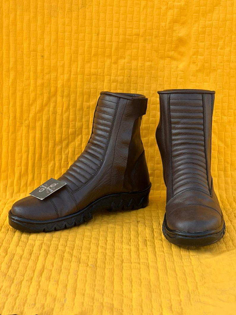 May include: A pair of brown leather ankle boots with a ridged design on the upper. The boots have a black sole and a small tag attached to one boot. The boots are displayed on a yellow textured surface.
