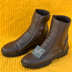 May include: A pair of brown leather boots with a ridged design on the upper shaft. The boots have a round toe and a black sole. The boots are displayed on a yellow background. A tag that says "Costume Replicas" is attached to one of the boots.