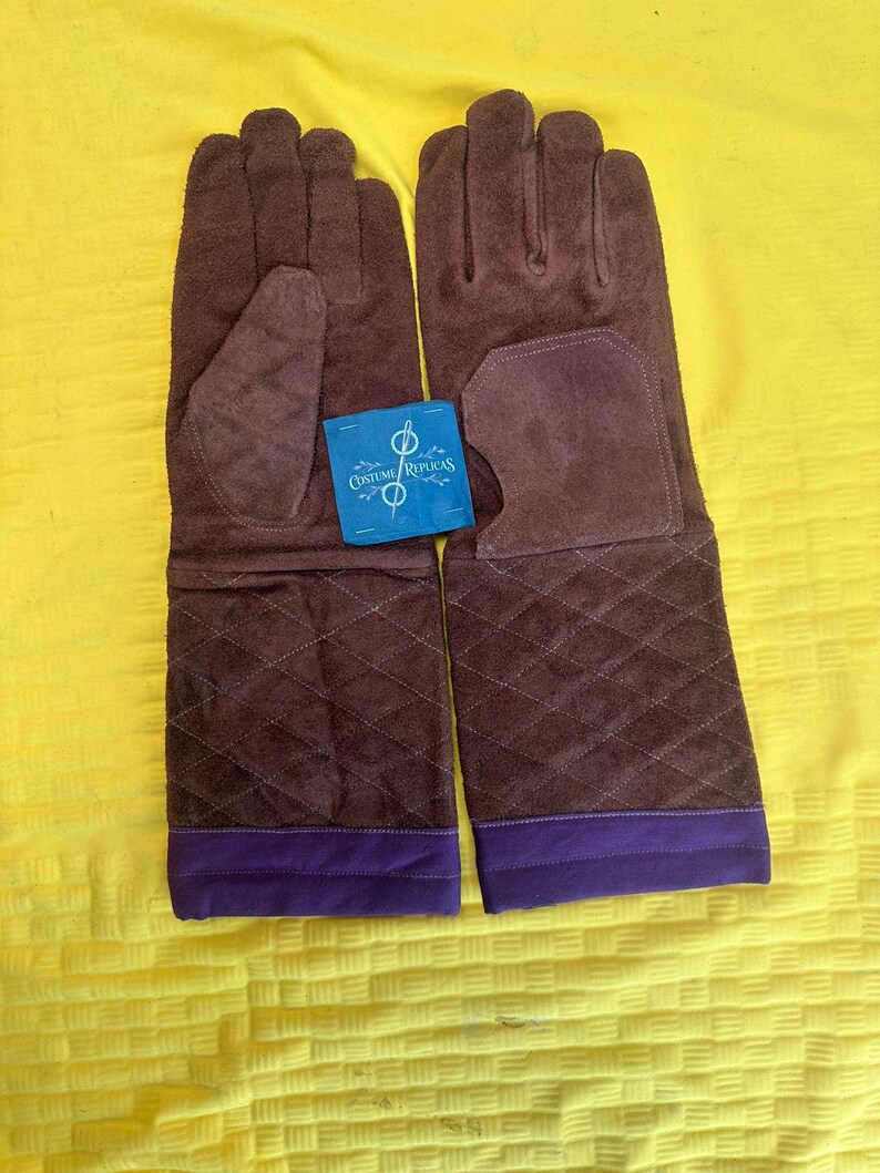 May include: A pair of brown suede gloves with quilted diamond stitching on the lower arm. The gloves have a purple band at the wrist and a blue tag that reads "Costume Replicas".