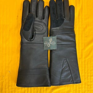 Può includere: Un paio di guanti in pelle nera con un design a polsino lungo. I guanti hanno una superficie strutturata e presentano cuciture rinforzate e una sezione imbottita sul dorso della mano. È attaccata una piccola etichetta con il testo "Costume Replicas".