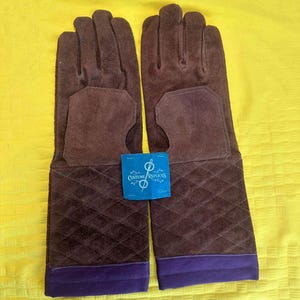 May include: A pair of brown suede gloves with long cuffs. The cuffs have a quilted pattern and a purple trim. A blue tag with the text "Costume Replicas" is between the gloves.
