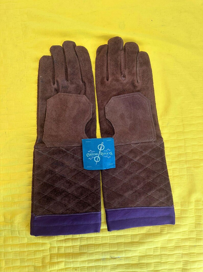 May include: A pair of brown suede gloves with long cuffs. The cuffs have a quilted pattern and a purple trim. A blue tag with the text "Costume Replicas" is between the gloves.