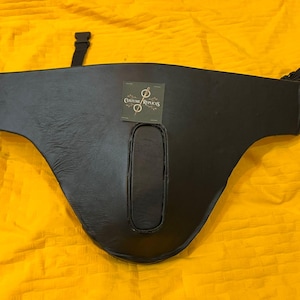 May include: A black leather chest armour piece with a curved shape and a central opening. It features two straps and a tag with a logo. The armour is displayed on a yellow surface.
