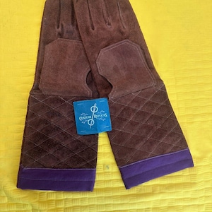 May include: A pair of brown suede gloves with a quilted pattern on the cuffs and a purple trim. The gloves have a cutout on the palm and a tag that reads "Costume Replicas".