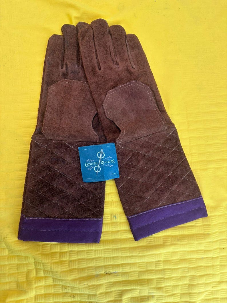 May include: A pair of brown suede gloves with a quilted pattern on the cuffs and a purple trim. The gloves have a cutout on the palm and a tag that reads "Costume Replicas".
