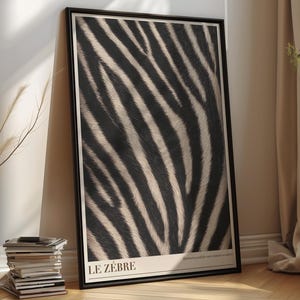 Op de afbeelding: Een rechthoekige ingelijste wandkunstprint met een contrastrijk zwart-wit zebrastreeppatroon. De print bevat de tekst LE ZÉBRE in de linkerbenedenhoek en kleinere tekst in de rechterbenedenhoek, alles geplaatst in een zwarte lijst.