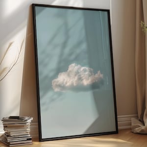 May include: Framed art print featuring a fluffy white cloud against a pale blue sky. The artwork is displayed in a black frame, leaning against a wall. The scene is illuminated by natural light, creating shadows.