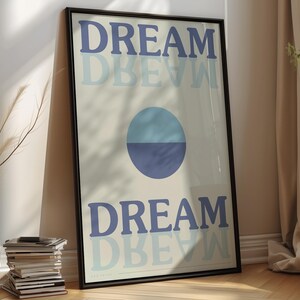 May include: Framed art print featuring the word "DREAM" in blue, with a shadow effect. A two-tone blue circle is centered. The print is in a black frame, leaning against a wall.