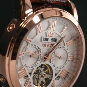 May include: Rose gold-toned wristwatch with a white face, featuring Roman numerals and a visible mechanical movement. The watch has a brown leather band and a crown design at the top. The watch face displays the day of the week and date.