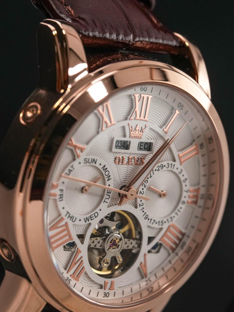 May include: Rose gold-toned wristwatch with a white face, featuring Roman numerals and a visible mechanical movement. The watch has a brown leather band and a crown design at the top. The watch face displays the day of the week and date.