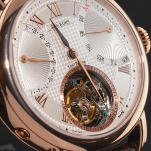 May include: Close-up of a rose gold wristwatch with a white dial. The watch features Roman numerals, a day-of-the-week display, and a tourbillon movement. The watch has a brown leather strap and the text "SIA-KORS" is visible.