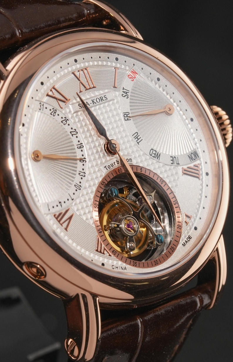 May include: Close-up of a rose gold wristwatch with a white dial. The watch features Roman numerals, a day-of-the-week display, and a tourbillon movement. The watch has a brown leather strap and the text "SIA-KORS" is visible.