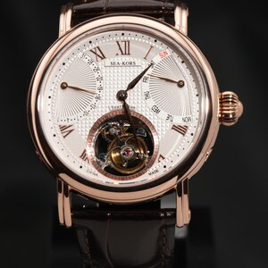 May include: A rose gold wristwatch with a white face, featuring Roman numerals and a visible tourbillon mechanism. The watch has a brown leather strap and the brand name "SEA-KORS" is visible. The watch face displays the days of the week and the date.