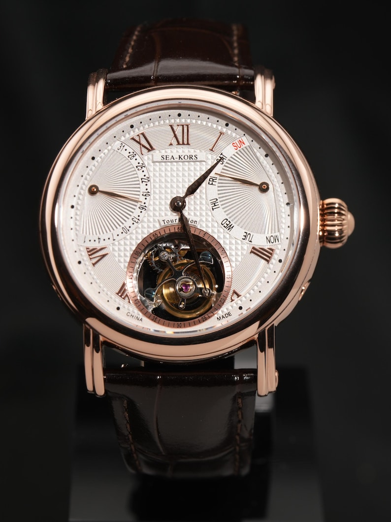 May include: A rose gold wristwatch with a white face, featuring Roman numerals and a visible tourbillon mechanism. The watch has a brown leather strap and the brand name "SEA-KORS" is visible. The watch face displays the days of the week and the date.