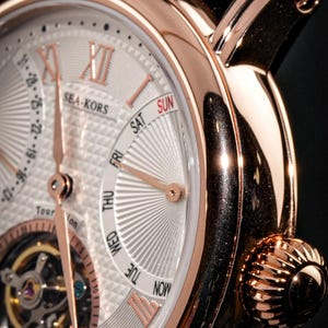 May include: Close-up of a rose gold wristwatch with a white face. The watch features Roman numerals, day and date indicators, and the brand name "SEA-KORS". The watch has a brown leather strap and a textured crown.