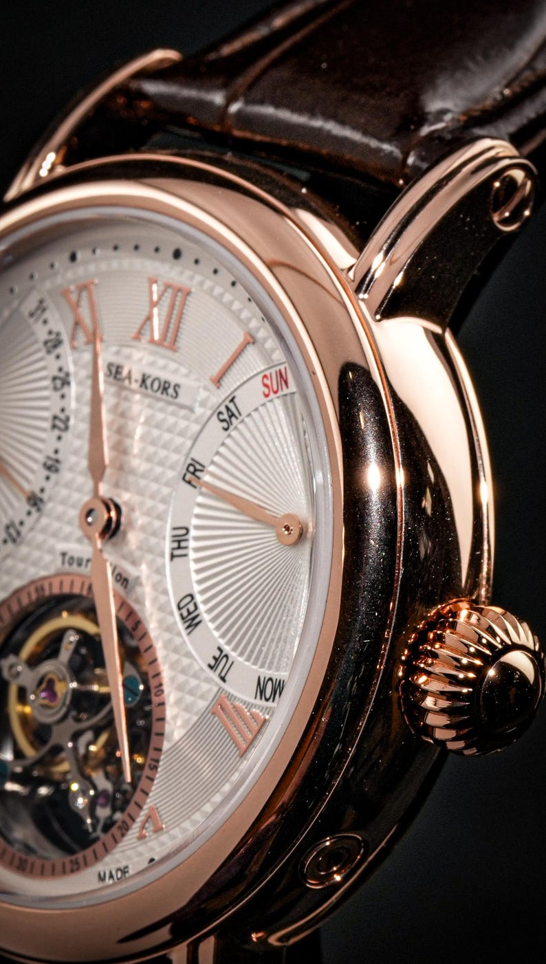 May include: Close-up of a rose gold wristwatch with a white face. The watch features Roman numerals, day and date indicators, and the brand name "SEA-KORS". The watch has a brown leather strap and a textured crown.