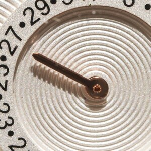 May include: Close-up of a watch face with a silver-colored background and a brown hand. The face has a circular design with concentric lines and black numbers marking the hours. The watch hand points towards the numbers.