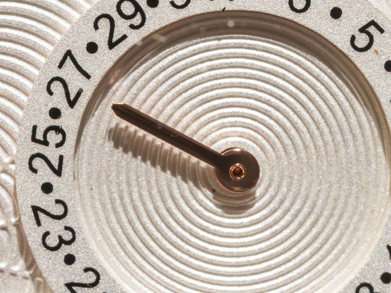 May include: Close-up of a watch face with a silver-colored background and a brown hand. The face has a circular design with concentric lines and black numbers marking the hours. The watch hand points towards the numbers.
