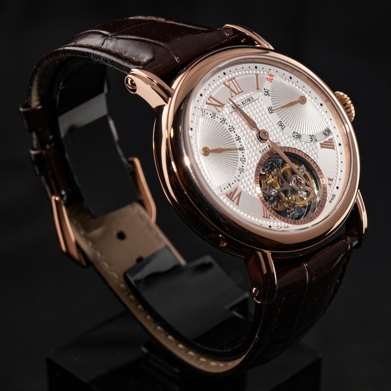 May include: A rose gold-toned wristwatch with a brown leather band. The watch face is white with Roman numerals and a visible mechanical movement. The watch displays the day of the week and date. The watch is displayed on a black stand.