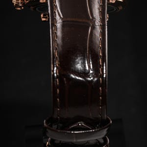 May include: Close-up of a luxury wristwatch with a brown leather band and a gold-colored case. The band has a textured, crocodile-like pattern. The watch's crown and buckle also have a gold finish. The watch is set against a dark background.