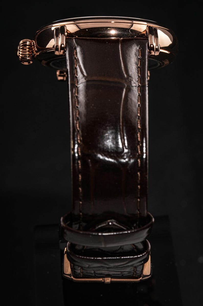 May include: Close-up of a luxury wristwatch with a brown leather band and a gold-colored case. The band has a textured, crocodile-like pattern. The watch's crown and buckle also have a gold finish. The watch is set against a dark background.
