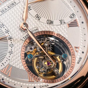 May include: Close-up of a luxury wristwatch with a silver face and rose gold accents. The watch features a visible tourbillon mechanism and day-of-the-week and date displays. The watch face has a textured pattern and Roman numeral hour markers.