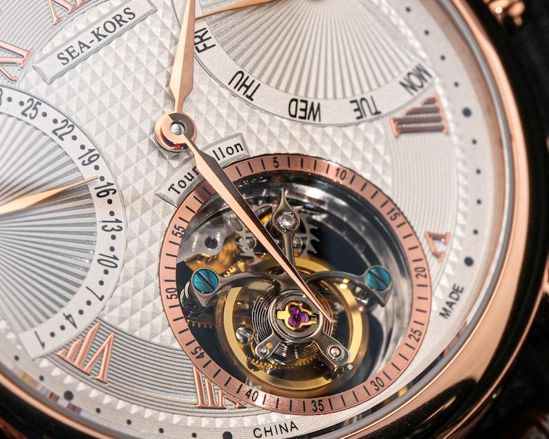 May include: Close-up of a luxury wristwatch with a silver face and rose gold accents. The watch features a visible tourbillon mechanism and day-of-the-week and date displays. The watch face has a textured pattern and Roman numeral hour markers.