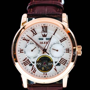 May include: A gold-toned OLEV watch with a white face, featuring Roman numeral hour markers and multiple sub-dials. The watch has a brown leather band and a visible inner mechanism. The text "OLEV" and "2034 DEC" are visible on the watch face.