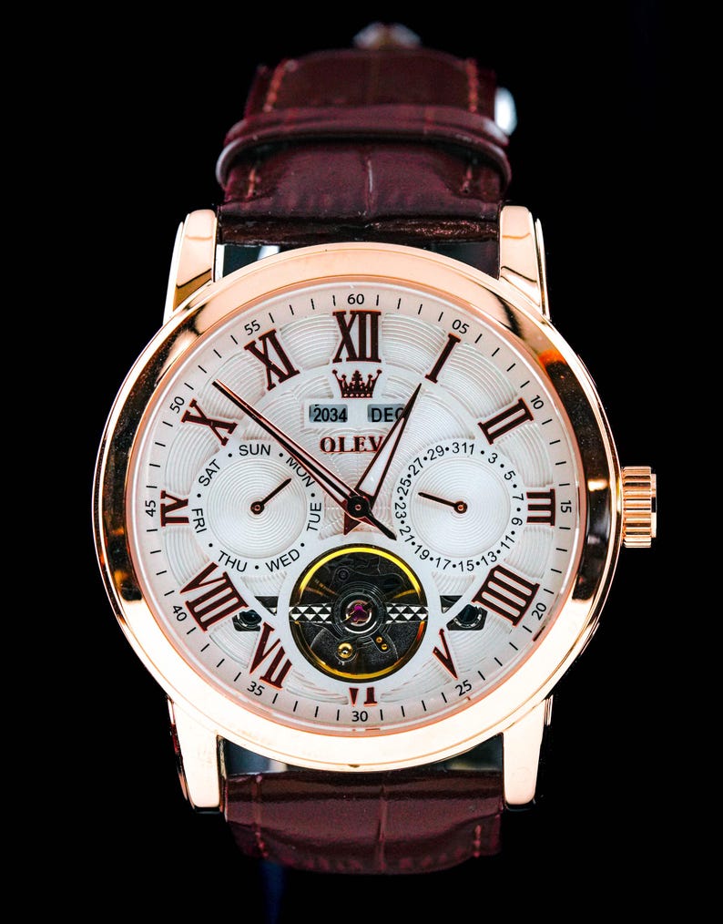 May include: A gold-toned OLEV watch with a white face, featuring Roman numeral hour markers and multiple sub-dials. The watch has a brown leather band and a visible inner mechanism. The text "OLEV" and "2034 DEC" are visible on the watch face.