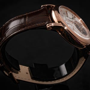May include: A rose gold wristwatch with a brown leather band. The watch face is white with Roman numerals and a visible mechanical movement. The band is a dark brown, textured leather. The watch is displayed against a black background.