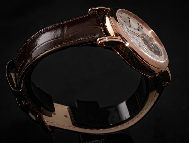 May include: A rose gold wristwatch with a brown leather band. The watch face is white with Roman numerals and a visible mechanical movement. The band is a dark brown, textured leather. The watch is displayed against a black background.