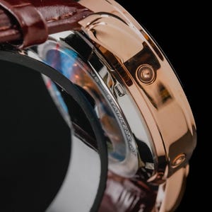 May include: Close-up of a rose gold wristwatch with a brown leather band. The watch face is partially visible, with a stainless steel casing and a detailed dial. The watch has a classic design.
