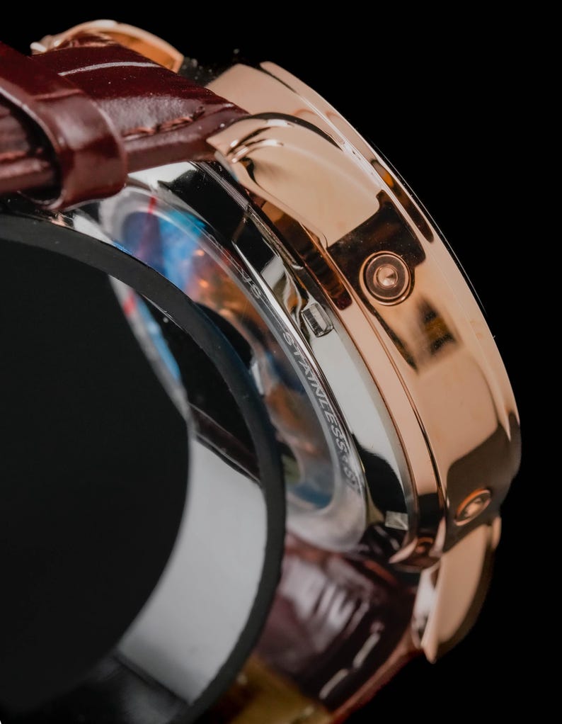 May include: Close-up of a rose gold wristwatch with a brown leather band. The watch face is partially visible, with a stainless steel casing and a detailed dial. The watch has a classic design.