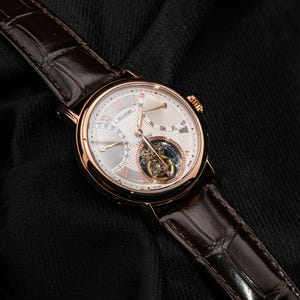 May include: A luxury wristwatch with a rose gold case and a white dial. The watch features a brown leather strap and a visible tourbillon movement. The dial includes sub-dials and Roman numeral hour markers. The watch is displayed on a black surface.